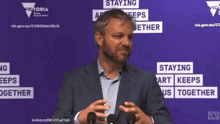 a man in a suit stands in front of a purple sign that says " staying apart keeps us together "