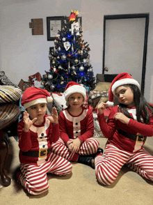 three children wearing santa hats and pajamas sit in front of a christmas tree