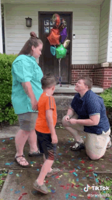 a man is kneeling down to propose to a woman and a boy in front of a house with balloons and confetti .