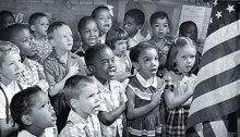 a group of children are singing the national anthem in a classroom while holding an american flag .