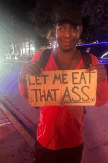 a man in a red shirt holds up a sign that says let me eat that ass