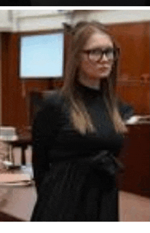 a woman in a black dress and glasses is standing with her arms crossed in a courtroom .