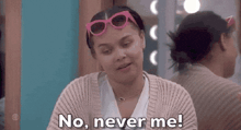 a woman wearing pink sunglasses and a sweater is sitting in front of a mirror and saying `` no , never me ! ''