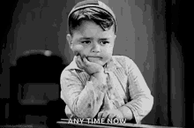 a black and white photo of a little boy sitting at a desk with his hand on his chin and the words `` any time now '' .