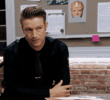 a man in a black shirt and tie sits at a desk in front of a bulletin board that says carisibot on it