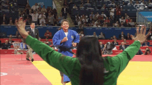 a woman in a green shirt stands in front of a judo match with the number 103 on the wall behind her