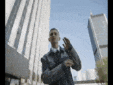 a man in a denim jacket and tie is standing in front of a tall building in a city .