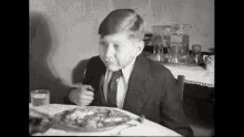 a young boy in a suit and tie is sitting at a table with a plate of food and a glass of water .