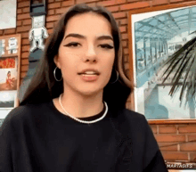 a woman wearing a pearl necklace and hoop earrings is standing in front of a brick wall and a picture .