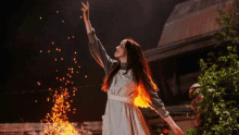 a woman in a white dress is standing in front of a fire reaching for something .