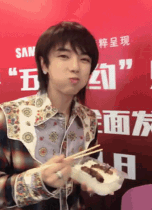 a young man is eating food with chopsticks in front of a red wall with chinese writing on it .