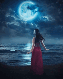 a woman in a red dress is standing on a beach at night looking at the moon .