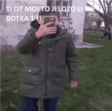 a young man in a green jacket is taking a selfie with his phone .