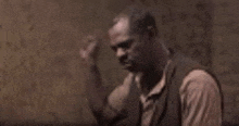 a man in a brown vest is sitting in a dark room with his hands on his head .