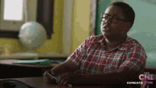 a boy in a plaid shirt sits at a desk in front of a sign that says " the chi " on it