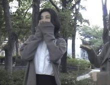 a woman is covering her face with her sweater while standing in a park with trees in the background .