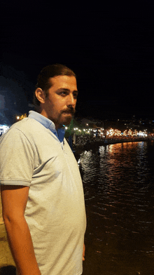 a man in a light blue shirt stands in front of a body of water at night