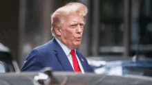 donald trump wearing a blue suit and red tie stands in front of a car