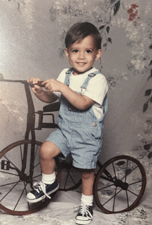 a young boy sitting on a tricycle wearing blue overalls