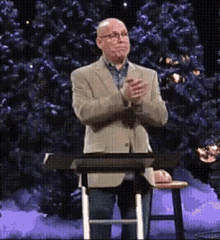 a man in a suit and glasses stands at a podium with his hands folded in front of a christmas tree