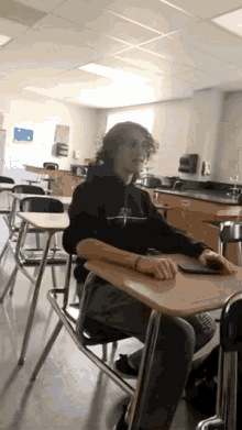 a young man sits at a desk in a classroom wearing a black sweatshirt with a cross on it ..