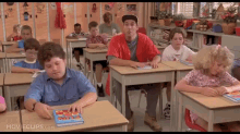 a group of children are sitting at their desks in a classroom with a man in a red shirt standing behind them .