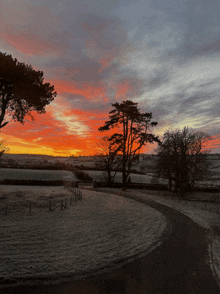 a sunset over a snowy field with a fence and trees in the foreground