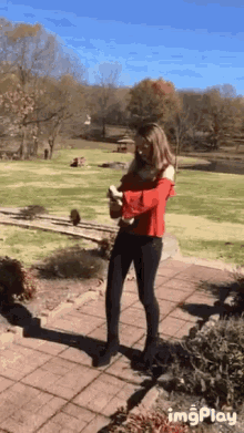 a woman in a red top and black pants is standing on a brick walkway in front of a field ..