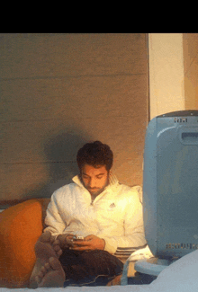 a man in an adidas jacket sits on a couch looking at his phone