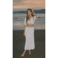 a woman in a white skirt and crop top stands on the beach