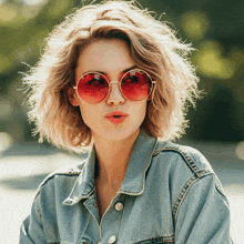 a woman wearing red sunglasses and a denim jacket blows a kiss