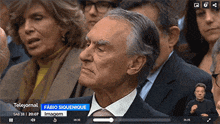 a man in a suit and tie is sitting in front of a screen that says telejornal fabio siquenique imagem