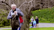 a man with a backpack is standing next to a tree in a park with a family in the background .