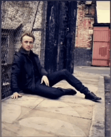 a man in a black jacket sits on the sidewalk with his legs crossed