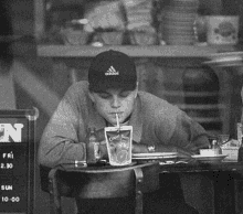 a man wearing an adidas hat sits at a table drinking capri sun juice