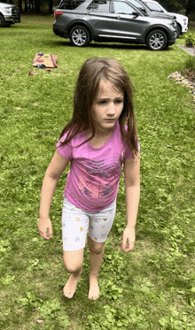 a little girl in a purple shirt and white shorts is standing in the grass