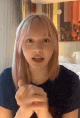 a woman with pink hair is sitting in a room with her hands folded and talking .