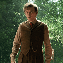 a man wearing glasses and a vest stands in front of a forest