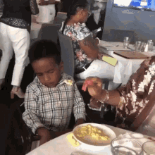 a little boy is sitting at a table with a bowl of food and a woman holding a piece of meat .