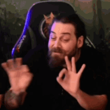 a man with a beard is sitting in a gaming chair and making an ok sign with his hands .