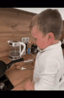 a little boy is standing in front of a stove in a kitchen looking at a frying pan .