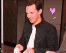 a man in a black jacket is sitting at a table with a cup of coffee in his hand .