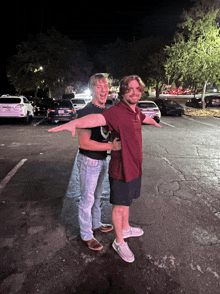 two men are posing for a picture in a parking lot at night with cars parked in the background