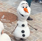 a stuffed snowman with an orange nose is standing on a brick sidewalk with a manhole cover in the background .