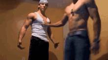 two muscular men are standing next to each other in a room . one of the men is wearing a hat .