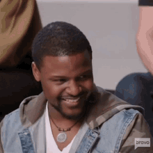 a man wearing a denim jacket and a necklace is smiling for the camera while sitting on a couch .