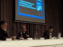 three men sit at a table with microphones in front of a screen that says grazie