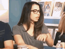 a woman wearing glasses sits at a table in front of a shelf with a girl 's magazine on it