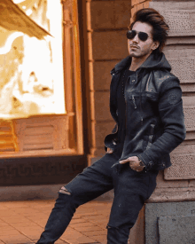 a man leaning against a wall wearing sunglasses and a leather jacket