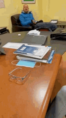 a man in a blue jacket is sitting at a desk with a stack of papers and a mask on it .